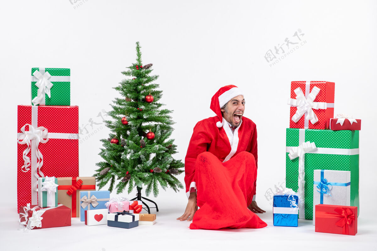 圣诞树滑稽感性的年轻人打扮成圣诞老人 拿着礼物和装饰过的圣诞树坐在白色背景的地上礼物礼物圣诞老人