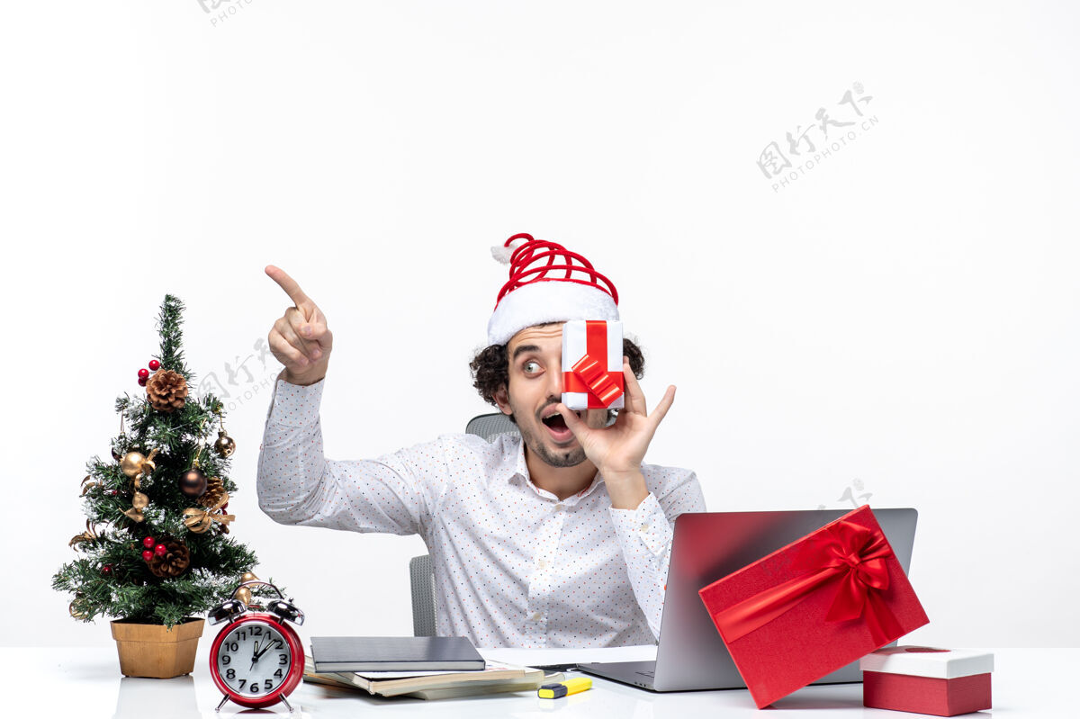 人圣诞节心情惊讶的商人与圣诞老人的帽子举起他的礼物 他的脸 并指出白色背景上的东西礼物孩子微笑