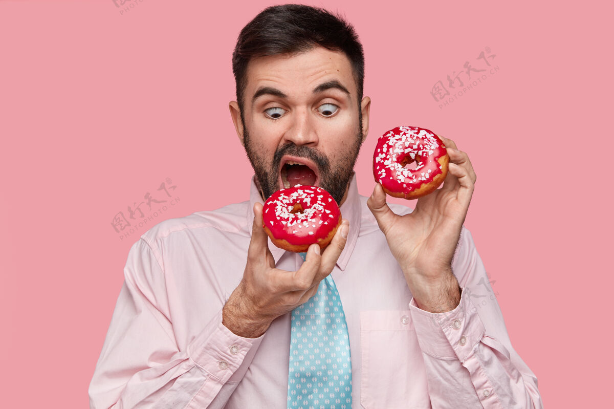 味道饥肠辘辘的帅哥咬着红甜甜圈 穿着正式衬衫打着领带 张大嘴巴饥饿咬甜点