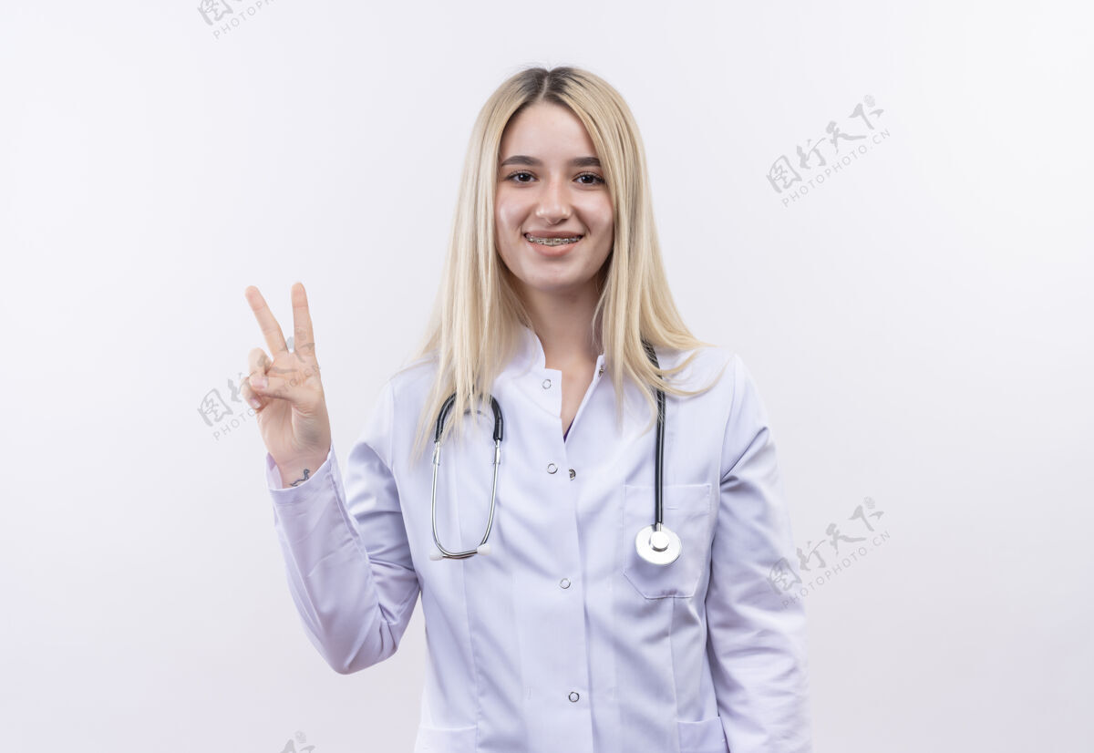 穿着微笑的医生 年轻的金发女孩 戴着听诊器 穿着医用长袍 戴着牙套 在孤立的白色背景上展示着和平的姿态医疗听诊器长袍