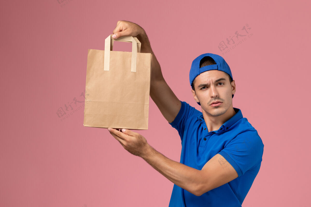 包裹正面图：身穿蓝色制服和斗篷的年轻男性快递员 手上拿着快递纸包裹 站在粉红色的墙上手成人微笑