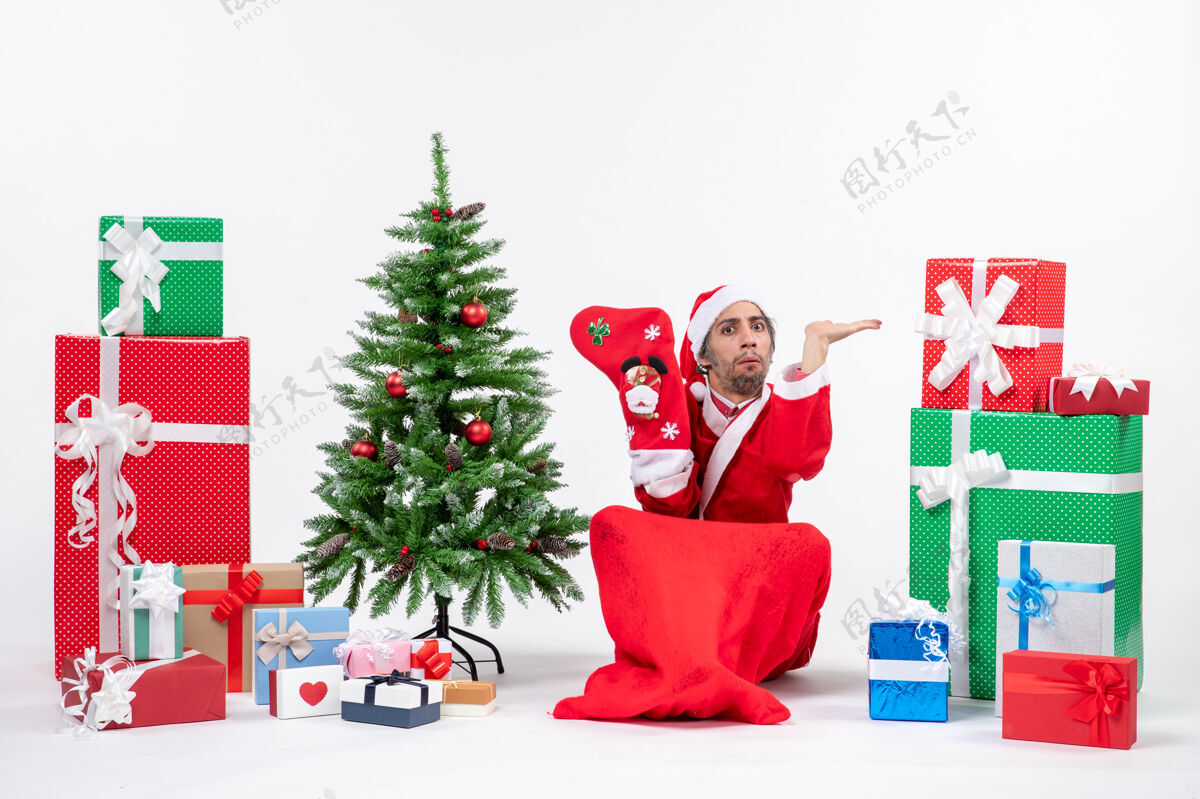 礼物严肃的圣诞老人坐在地上 穿着圣诞袜 靠近礼物和装饰在白色背景上的新年树严肃袜子坐