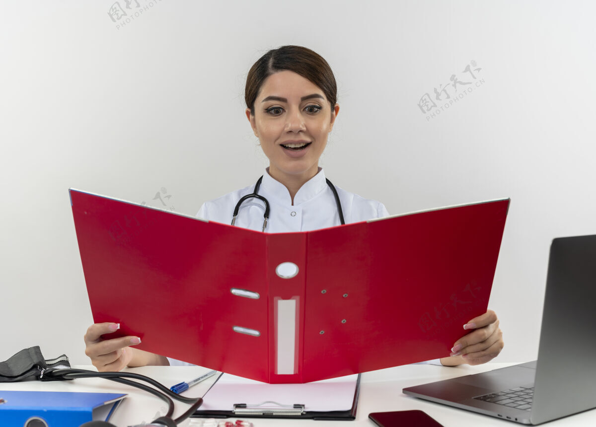 穿着高兴的年轻女医生穿着医用长袍带听诊器坐在办公桌上用医疗工具拿着电脑看文件夹和复印空间坐着办公桌女