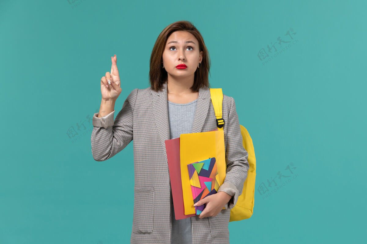 学生身穿灰色夹克 背着黄色背包 手拿文件和抄写本的女学生正对着蓝色的墙壁Female保持夹克