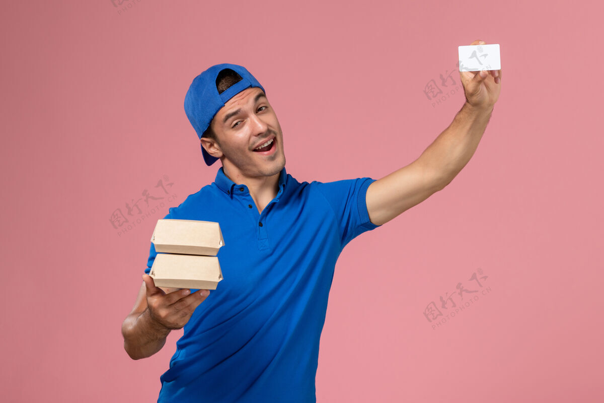 年轻正面图：身穿蓝色制服披肩的年轻男性快递员拿着小包裹 浅粉色墙上有卡片正面人持有