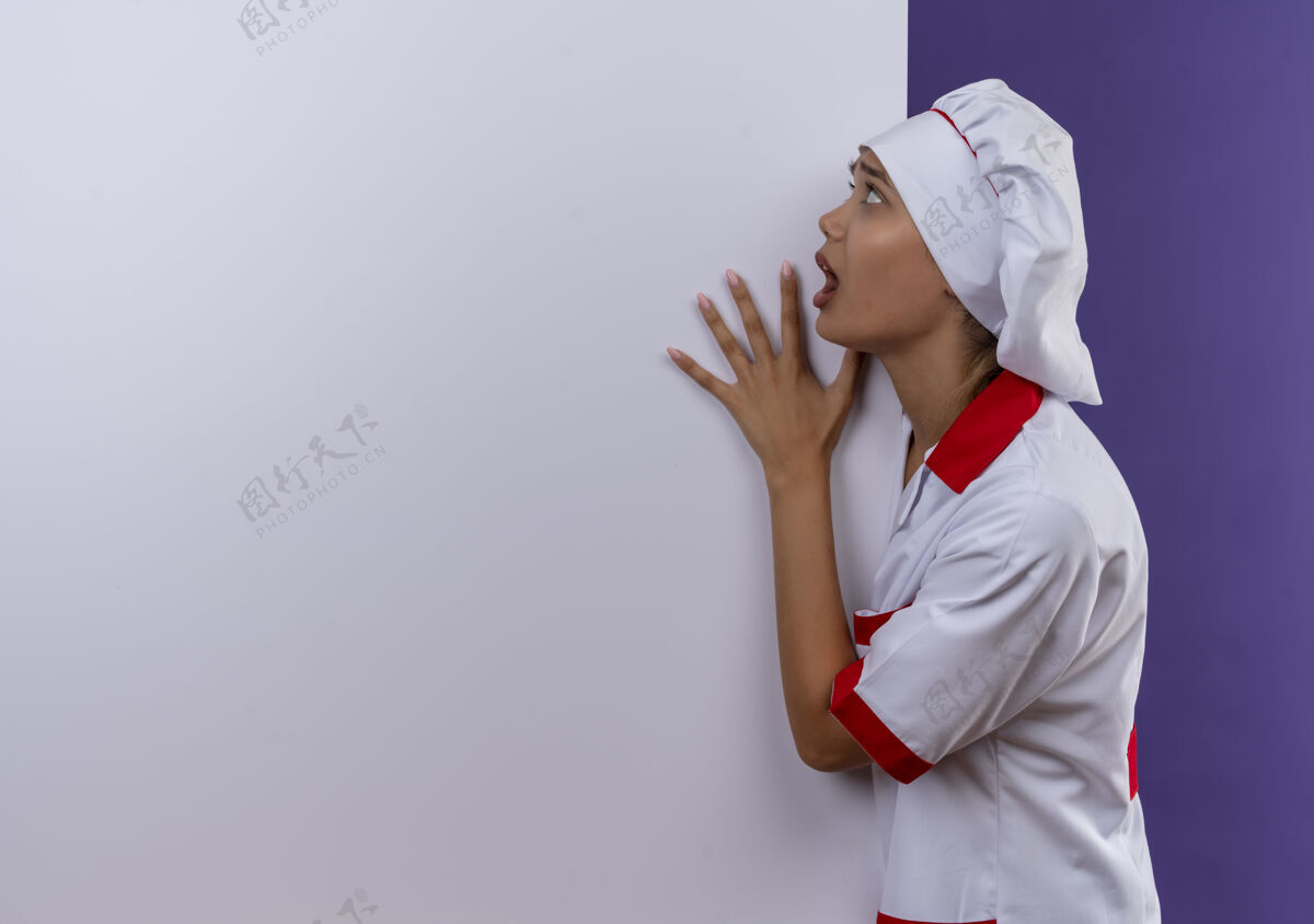 厨师害怕的年轻厨师女穿着厨师制服拿着白色墙壁在孤立的背景与复制空间拿着厨师制服