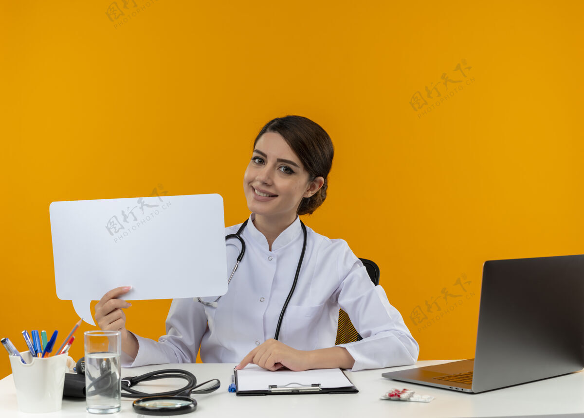 工作微笑的年轻女医生穿着医用长袍 手持听诊器 坐在办公桌旁 拿着医疗工具 拿着聊天泡泡 隔离黄色背景长袍医生穿