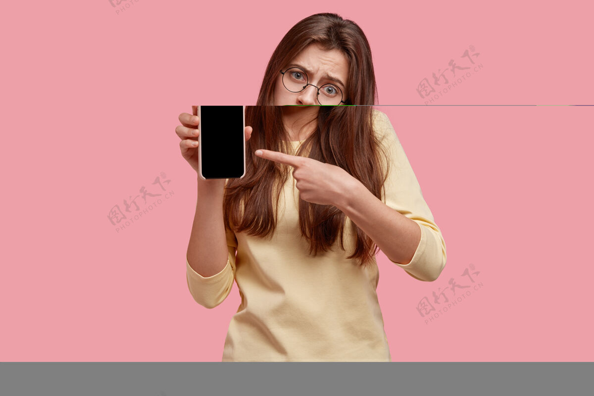 显示心烦意乱的垂头丧气的女人掏着下唇 指着现代的小玩意儿 在屏幕上显示你的文字 不喜欢它的工作方式 戴着圆眼镜智能手机通讯空白