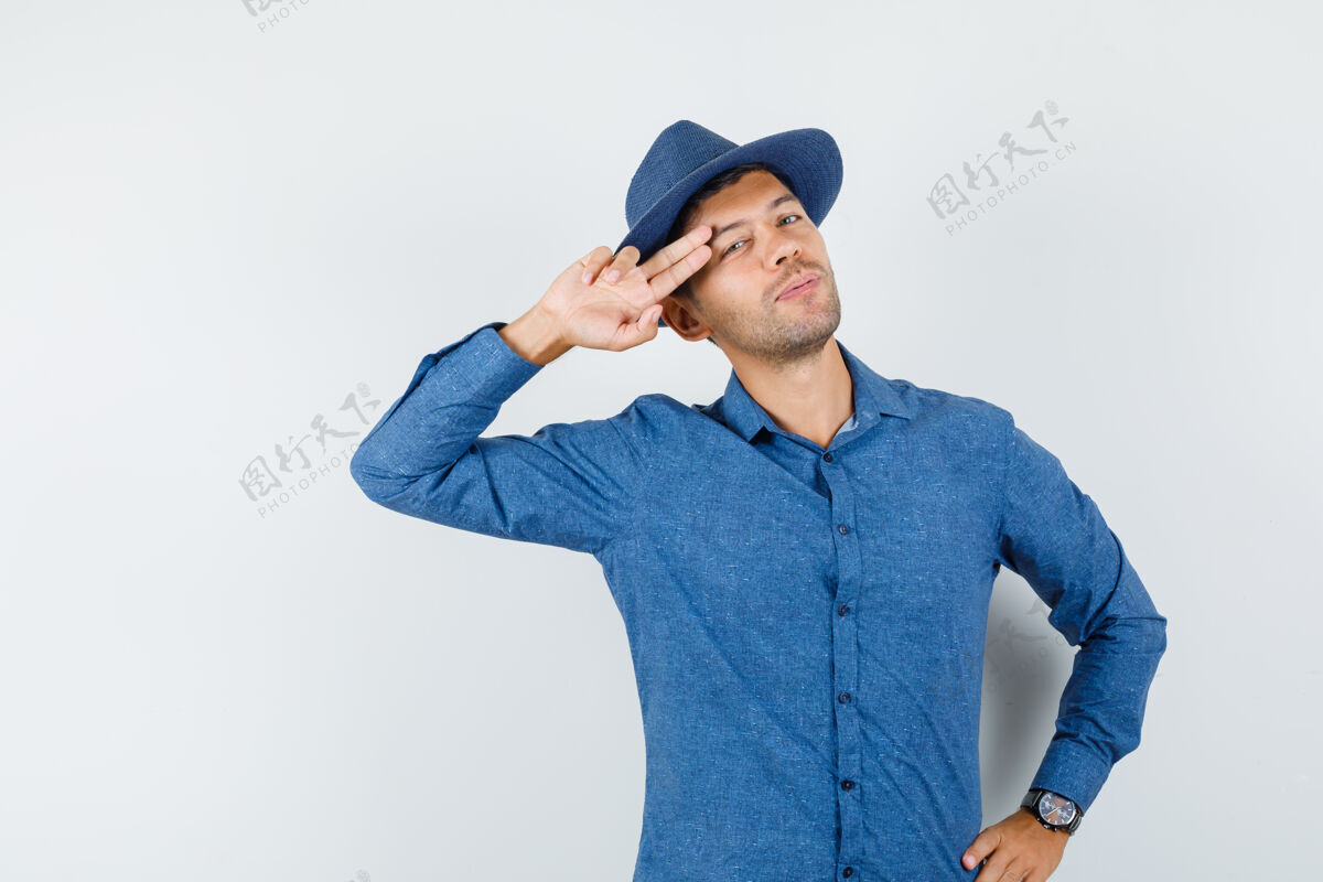 表情穿着蓝色衬衫 戴着帽子的年轻男子 两个手指放在太阳穴附近 看上去很酷 正面视图肖像自信站着