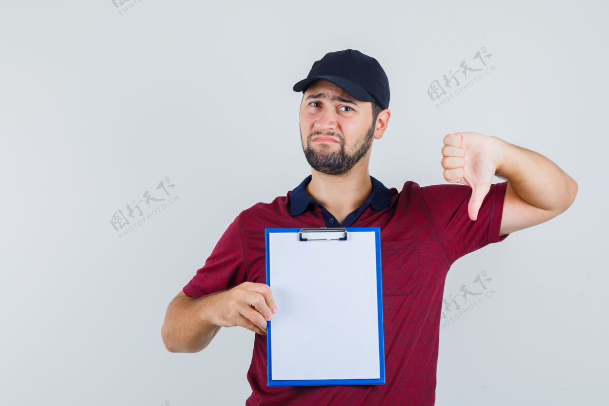 下身穿红色t恤 戴着黑色帽子的年轻男子拿着笔记本 大拇指朝下 看上去很悲伤 正面视图自信休闲笔记本