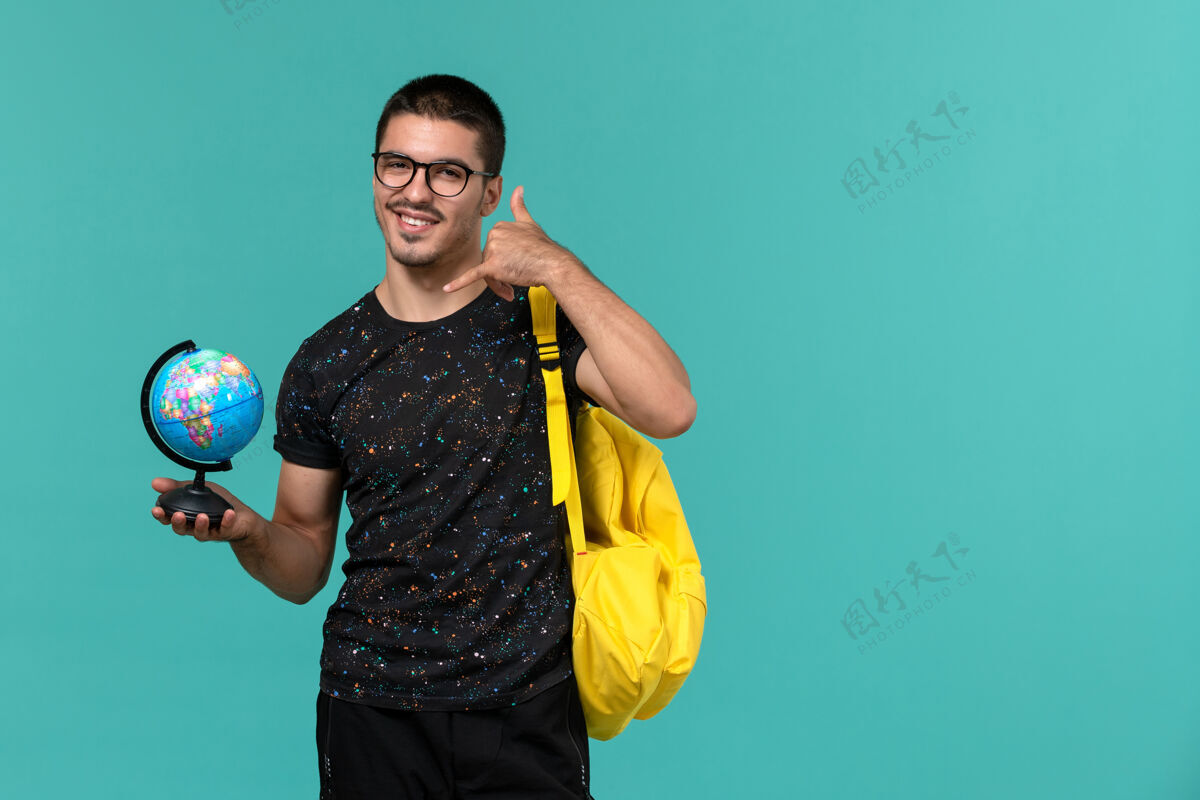 肖像身穿深色t恤黄色背包的男学生正面图蓝色墙上挂着微笑的小地球仪人背包人