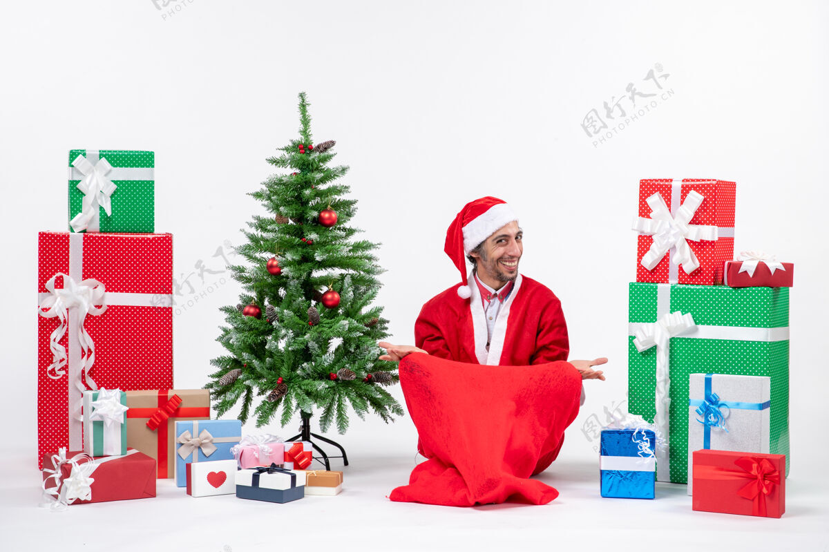成人满意的年轻人打扮成圣诞老人与礼物和装饰圣诞树坐在地上的白色背景装饰年轻人微笑