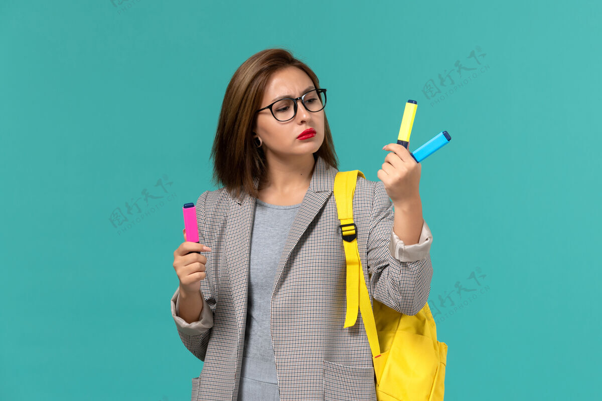 灯光身穿灰色夹克 背着黄色背包 拿着毛毡笔的女学生在浅蓝色墙上的正面照片刷子学院学生
