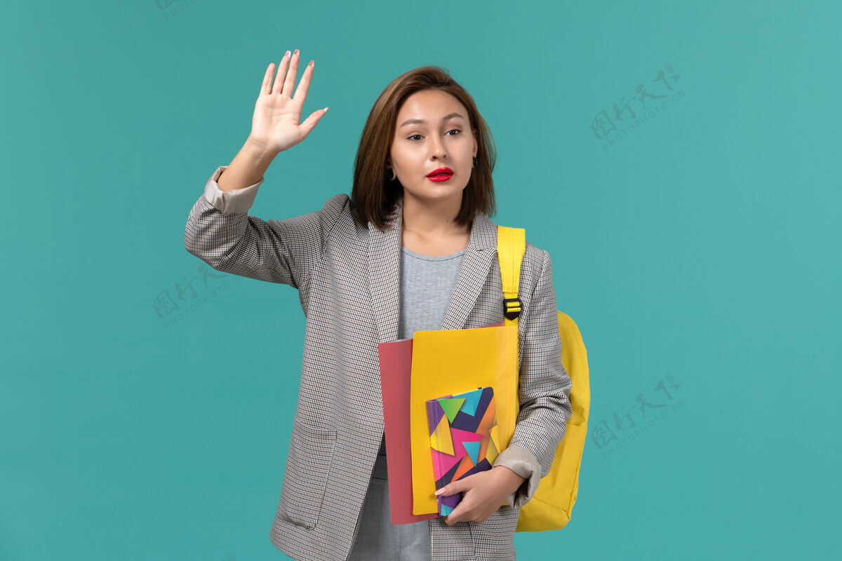学校身穿灰色夹克 背着黄色背包 拿着文件和文案的女学生在蓝色墙上挥舞的正面照片女学生背包大学