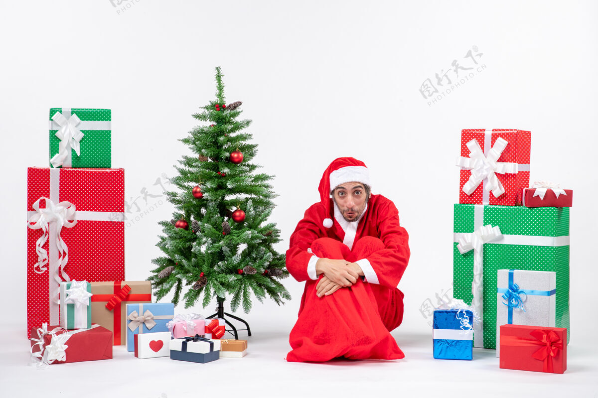 微笑困惑的年轻人打扮成圣诞老人 拿着礼物和装饰过的圣诞树坐在地上的白色背景上礼物年轻人装饰