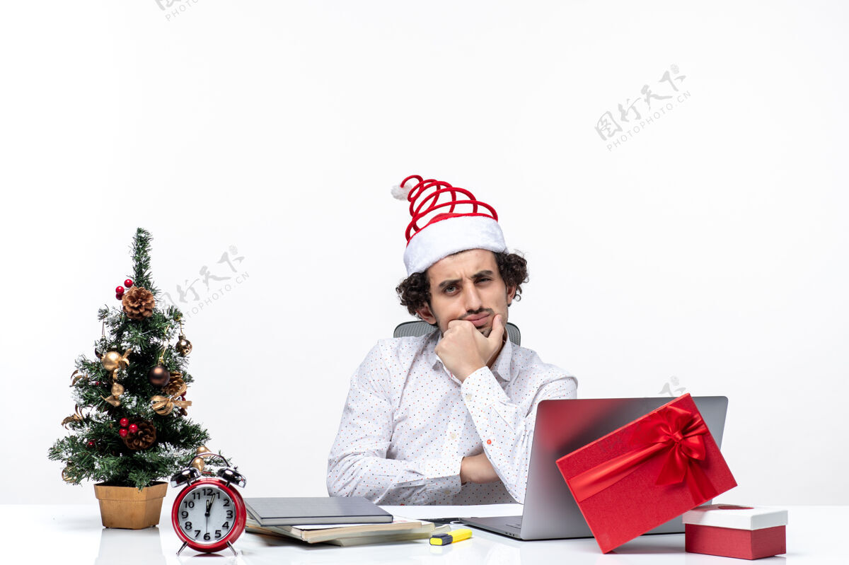 工作困惑的年轻商人戴着滑稽的圣诞老人帽 在办公室里思考着白色背景上的东西微笑年轻人