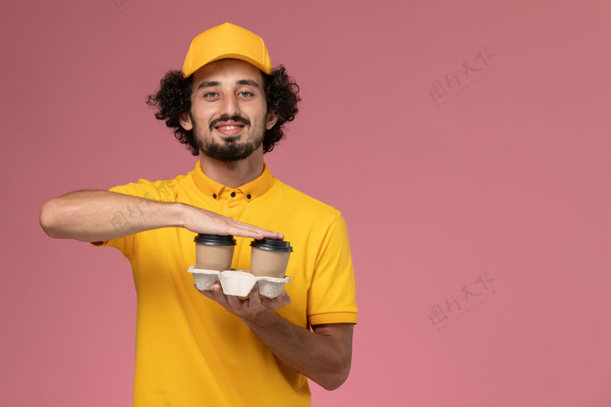 男正面图：身穿黄色制服和披风的男性信使手持棕色咖啡杯 微笑着站在粉红色的墙上人微笑咖啡