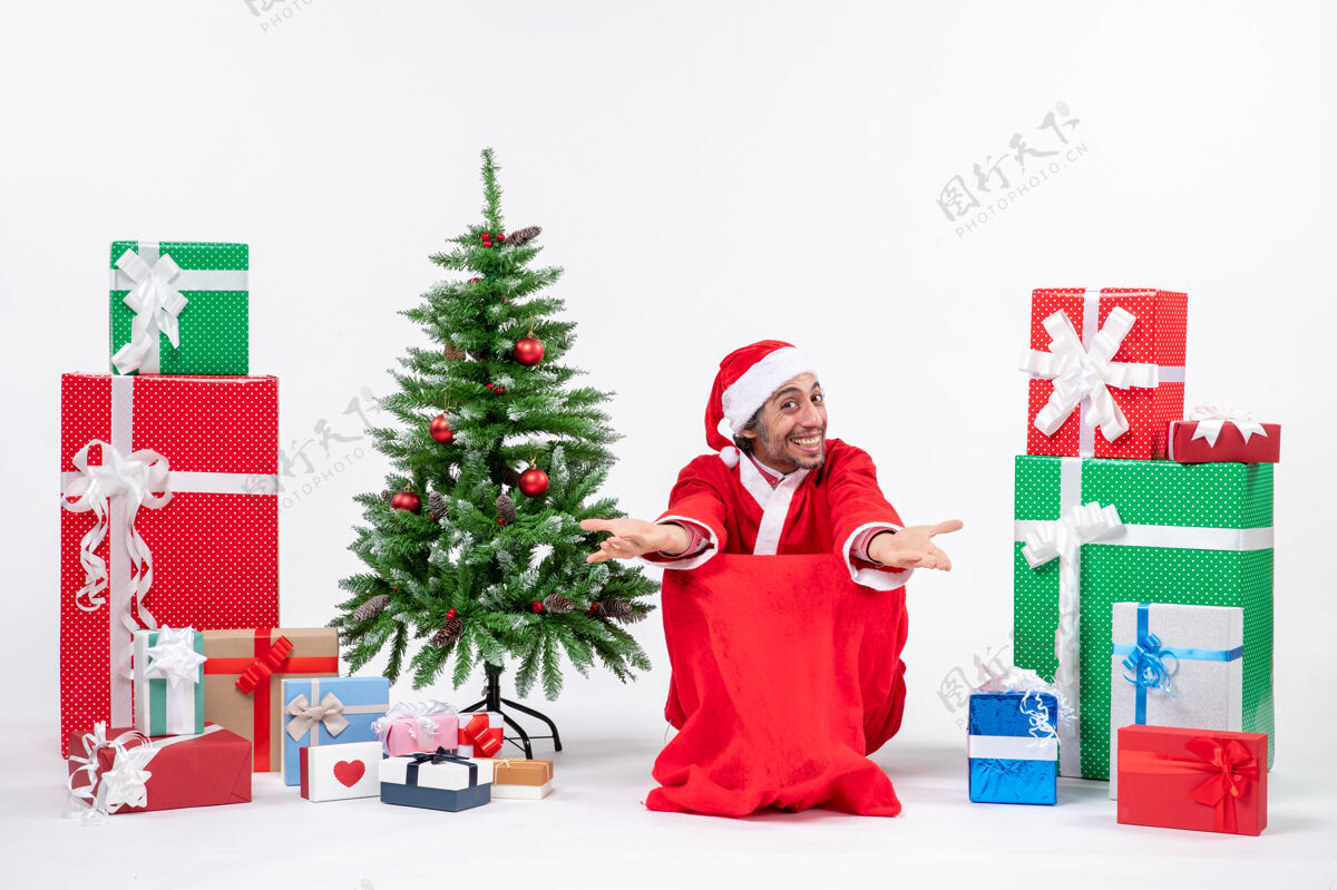人情绪激动的年轻人打扮成圣诞老人与礼物和装饰圣诞树欢迎有人在白色背景装饰打扮礼物