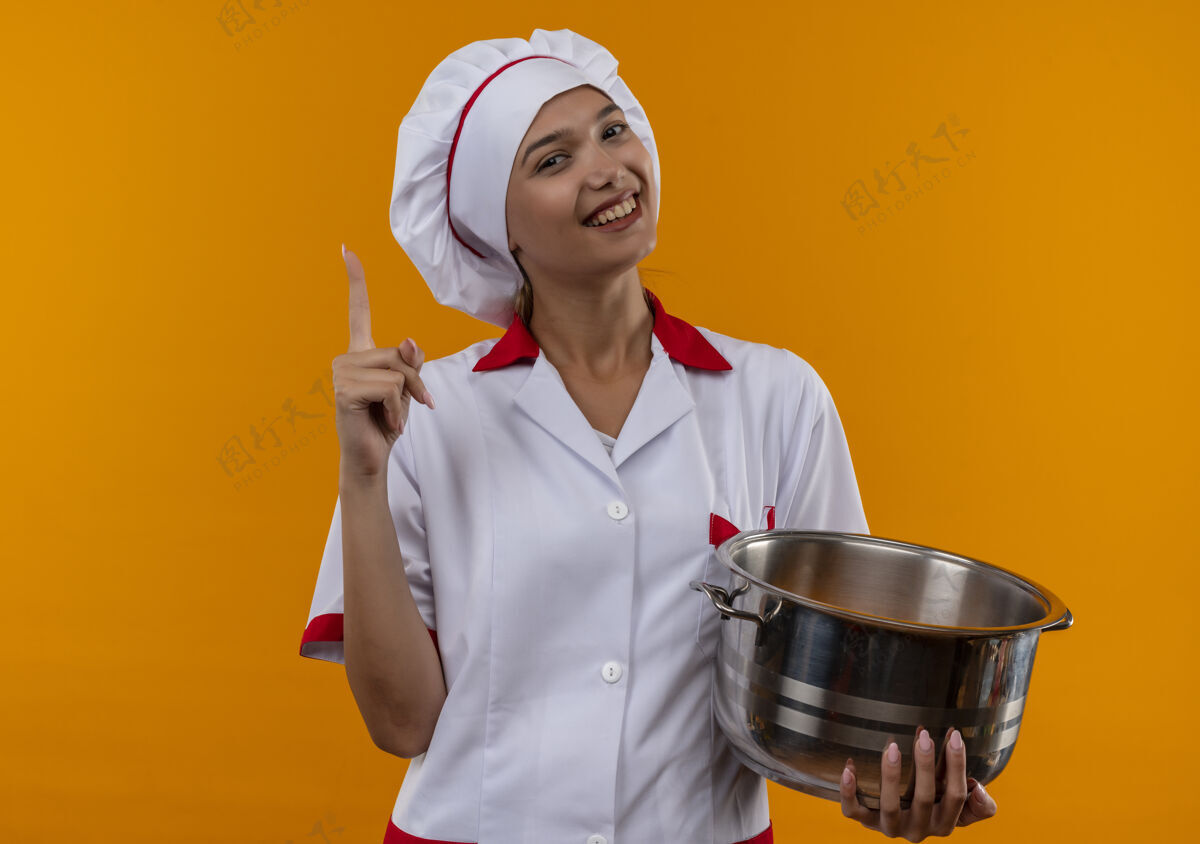 点微笑的年轻厨师女身穿厨师制服拿着平底锅手指向上在孤立的橙色背景与复制空间向上厨师女性