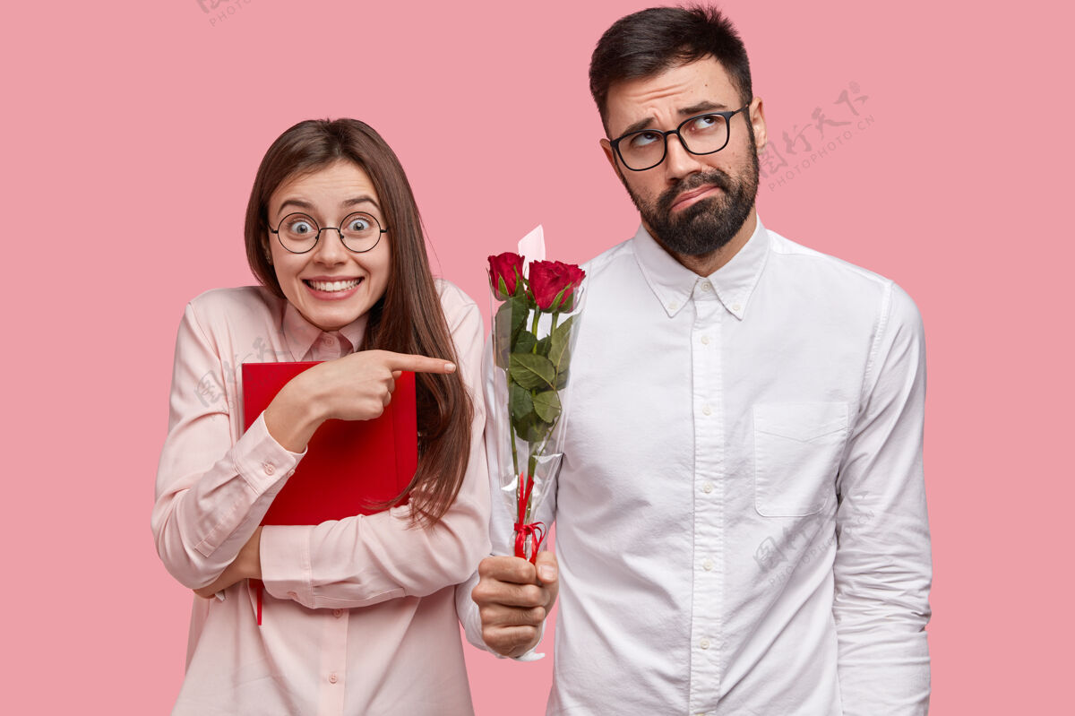 衬衫积极的欧洲年轻女子拿着红色课本 指着尴尬的白衬衫胡子男人 他觉得害羞 手里拿着漂亮的花束 有爱情故事姿势浪漫男性