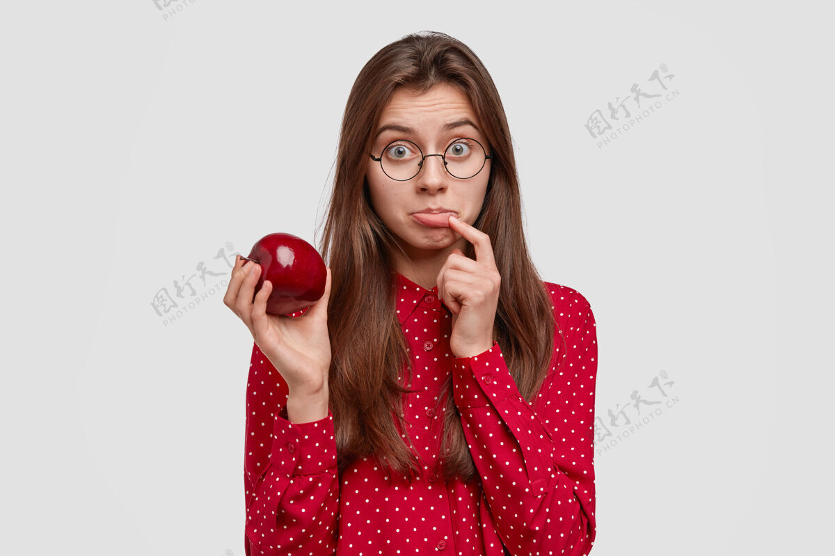 有机可悲可爱的女素食者手持新鲜的红苹果 包着下唇 保持健康的饮食 吃水果 留着长长的直发 穿着圆领上衣独自关心保健