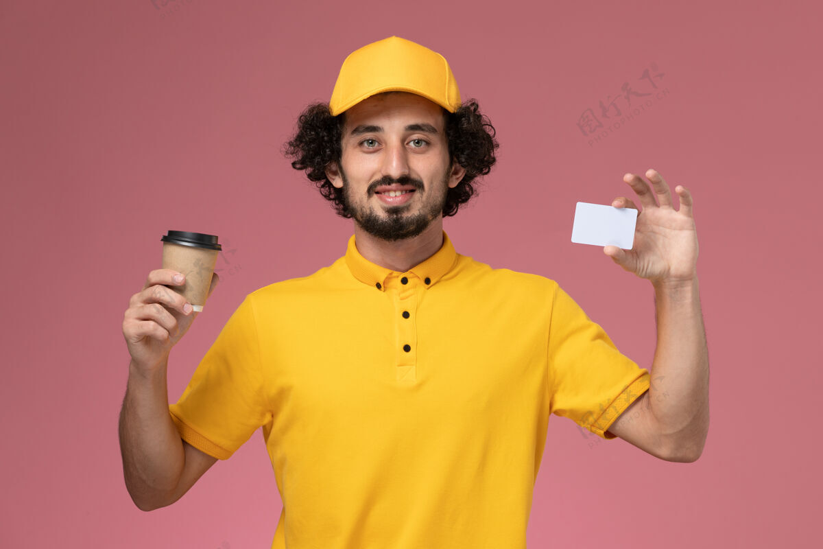 男快递正面图：身穿黄色制服 披风的男性快递员手持咖啡杯和粉色墙上的卡片快递专业服务