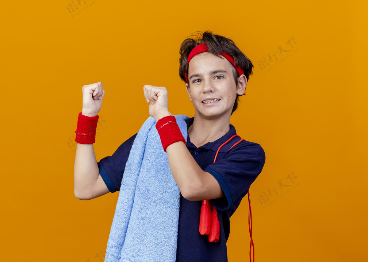 青少年面带微笑的年轻帅气的运动男孩戴着头带和腕带 肩上搭着毛巾和跳绳 指指点点身后看肩膀橙色