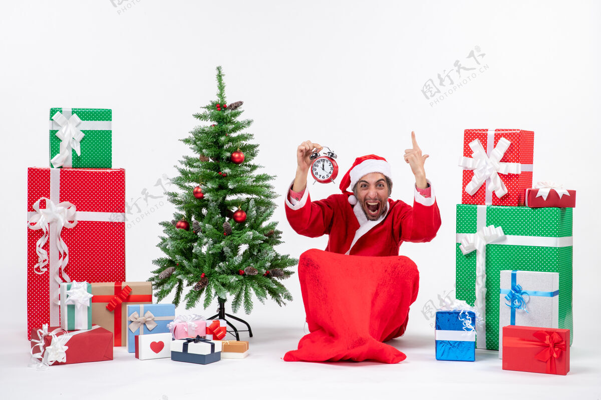 坐着紧张兴奋的圣诞老人坐在地上 在礼物和装饰圣诞树旁的白色背景上升起闹钟紧张饲养圣诞老人