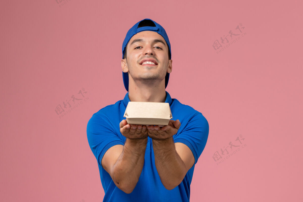 包裹正面图身穿蓝色制服披肩的年轻男性快递员拿着小快递食品包站在浅粉色的墙上送货送货制服