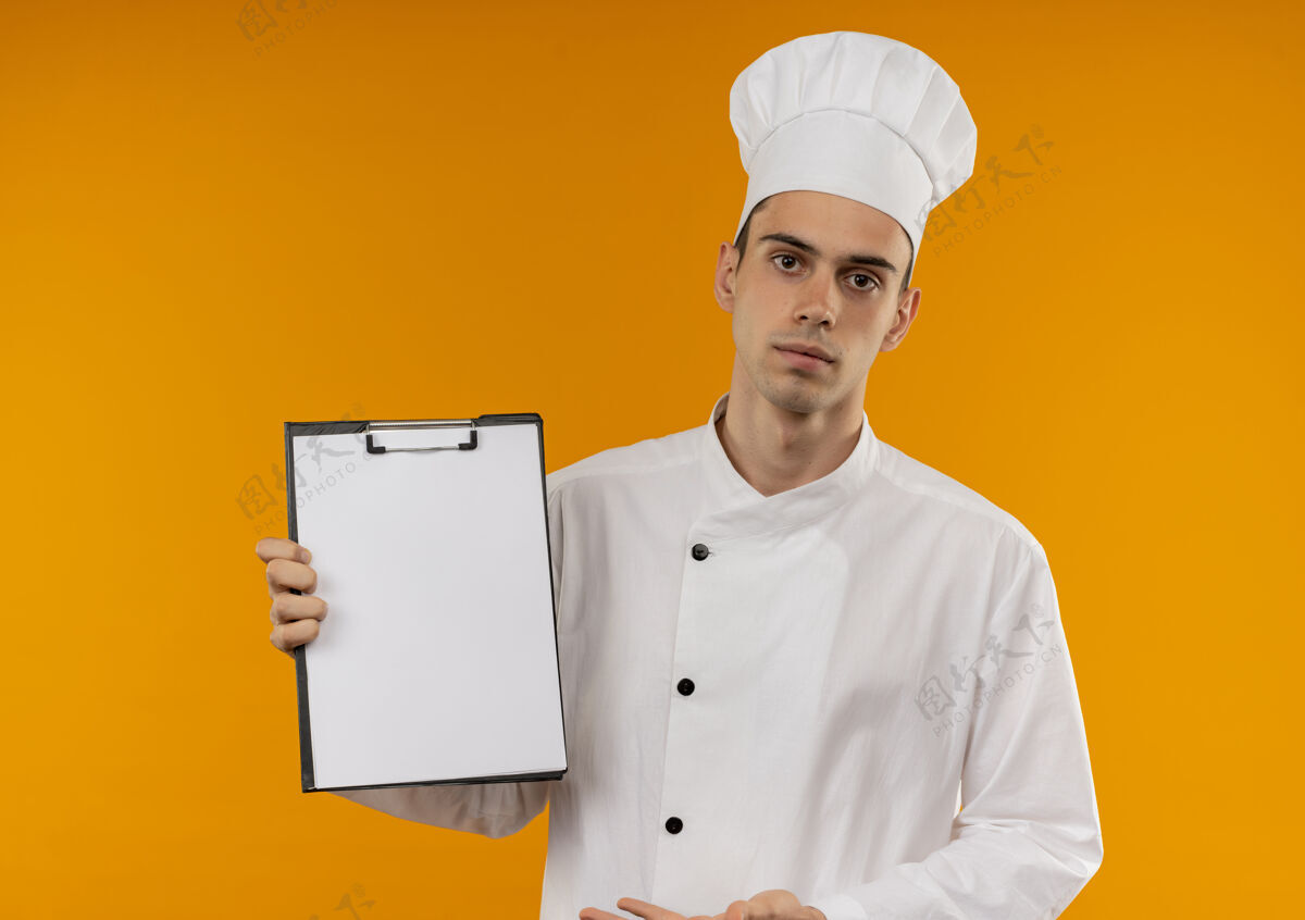 穿年轻的男酷穿着厨师制服显示剪贴板在他的手上孤立的黄色墙壁年轻酷厨师