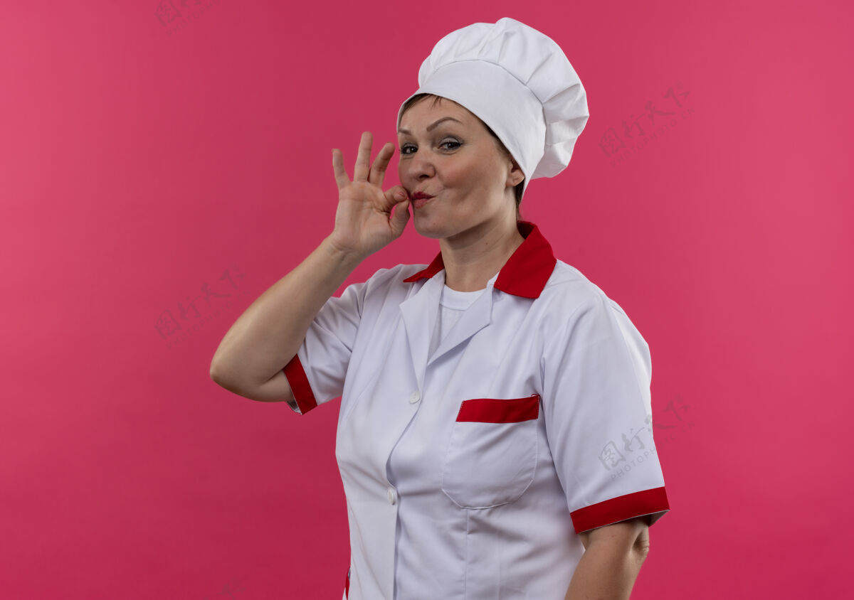 制服身着厨师制服的中年女厨师在隔离的粉色墙壁上展示着美味的手势 还有复制空间手势展示高兴