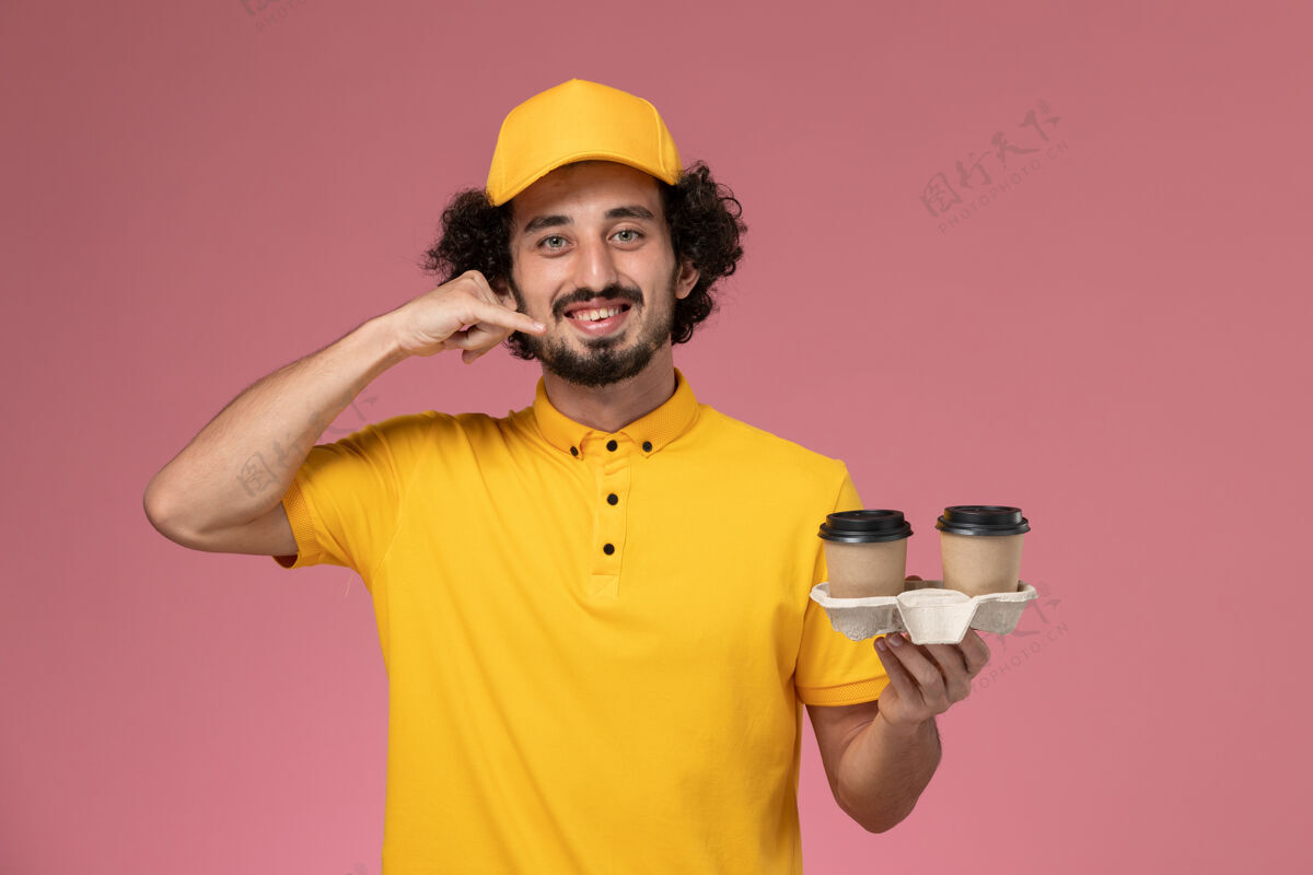 视野正面图：身穿黄色制服 披风的男性快递员在粉色墙上拿着送货咖啡杯男性快递员工作工人