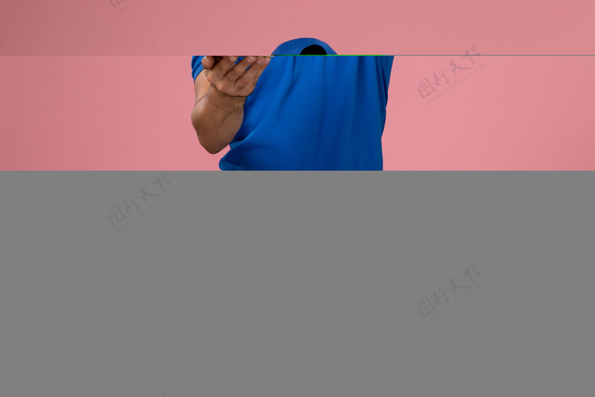 持有正面图：身穿蓝色制服披肩的年轻男性快递员在粉红色的墙上拿着小包裹服务男性消费者